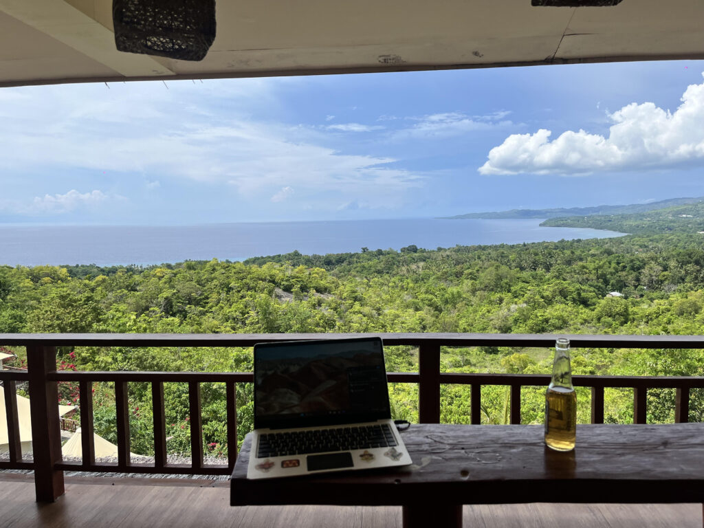 Writing a Newsletter! Remote game copywriters workspace overlooking a tropical coastline, with a laptop and beer on a balcony desk— freedom and creativity of working in the game industry from anywhere in Southeast Asia.